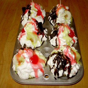 May include: A metal muffin tin holds six cupcake-shaped candles. Each candle is topped with white frosting, red and green drizzle, chocolate squares, and a cherry. A lit wick sits atop each candle.
