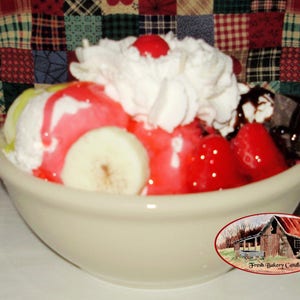 May include: A sundae in a cream-colored bowl, featuring scoops of ice cream, strawberries, a banana slice, whipped cream, and a cherry. The sundae is drizzled with red and chocolate syrup. The bowl has a Fresh Bakery Candles sticker.