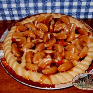 May include: A round apple pie candle with a golden crust and a filling of glazed apple slices. The pie sits in a silver foil pan. Small white candles are placed on top of the apple slices. The image includes a Fresh Bakery Candles logo.