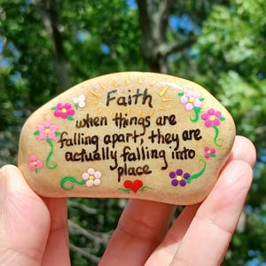May include: A smooth, tan-colored river stone painted with black text that reads, "Faith when things are falling apart, they are actually falling into place." The stone is decorated with small, colorful hand-painted flowers and a red heart.