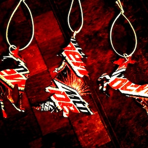 May include: Three red and white Christmas ornaments shaped like a reindeer, a tree, and another reindeer. The ornaments are made from recycled aluminum cans.