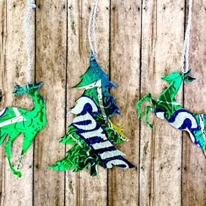 May include: Three Christmas ornaments made from recycled aluminum cans. The ornaments are shaped like a Christmas tree and two reindeer. The ornaments are green, blue, and silver.