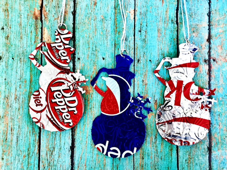 Puede incluir: Tres adornos de mu&ntilde;eco de nieve hechos con latas de aluminio recicladas. Los adornos est&aacute;n decorados con pintura roja, blanca y azul y tienen las palabras "Dr Pepper" y "Diet Dr Pepper" impresas.