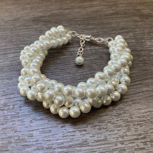 May include: A white pearl bracelet with a silver-toned clasp. The bracelet is made of multiple strands of small, round pearls woven together. The pearls have a lustrous sheen, and the bracelet is displayed on a wooden surface.