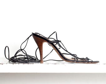 May include: A black strappy sandal with a high, wooden heel. The sandal features multiple thin straps that wrap around the ankle. The shoe is displayed on a white surface.