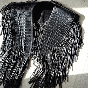 May include: Black leather vest with a crocodile texture and long fringe. The vest features a collar and a front closure. The fringe hangs from the shoulders and the bottom, creating a dramatic look.