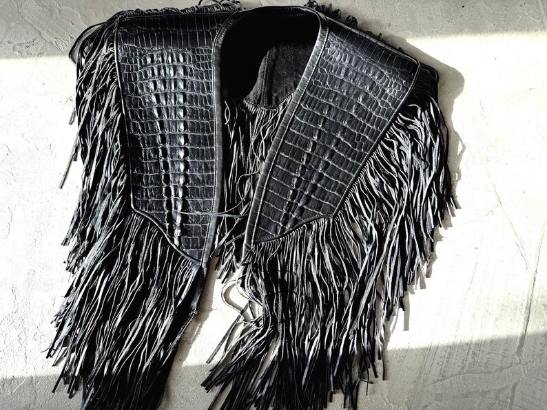 May include: Black leather vest with a crocodile texture and long fringe. The vest features a collar and a front closure. The fringe hangs from the shoulders and the bottom, creating a dramatic look.