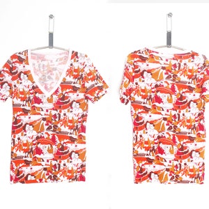 May include: A white short-sleeved t-shirt with a colourful, abstract print featuring orange, red, and brown hues. The print depicts a variety of figures and shapes.