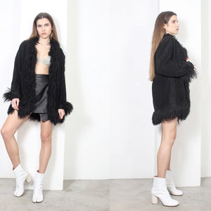 May include: A black suede jacket with a long, shaggy fur collar and trim. The jacket has a long, loose fit and is worn with a black mini skirt and white platform shoes.