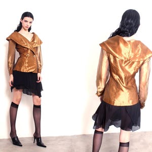 Könnte beinhalten: Goldfarbene Jacke mit weitem Kragen und langen Ärmeln, kombiniert mit einem schwarzen Rock und durchsichtigen schwarzen Kniestrümpfen. Das Model trägt schwarze Absätze. Die Jacke hat zwei goldene Knöpfe.