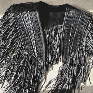 May include: Black leather jacket with fringe detailing. The jacket has a crocodile-textured pattern on the shoulders and collar. The fringe hangs from the shoulders and the bottom, creating a unique, stylish look.