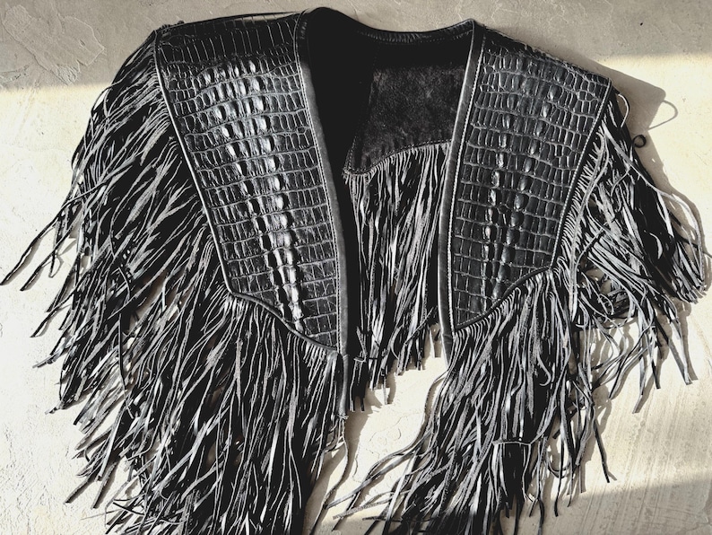 May include: Black leather jacket with fringe detailing. The jacket has a crocodile-textured pattern on the shoulders and collar. The fringe hangs from the shoulders and the bottom, creating a unique, stylish look.