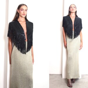 May include: A black fringed shawl with a V-neck design. The shawl is paired with a long, shimmering gold skirt. The outfit is set against a white background, creating a striking contrast. The overall look is stylish and elegant.