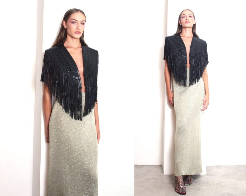May include: A black fringed shawl with a V-neck design. The shawl is paired with a long, shimmering gold skirt. The outfit is set against a white background, creating a striking contrast. The overall look is stylish and elegant.