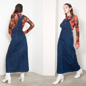 Op de afbeelding: Een lange, donkerblauwe denim overall jurk met een slab, zakken en knoopdetails. De jurk wordt gecombineerd met een top met lange mouwen met patroon en witte enkellaarsjes. De overall jurk is een klassiek en veelzijdig kledingstuk.