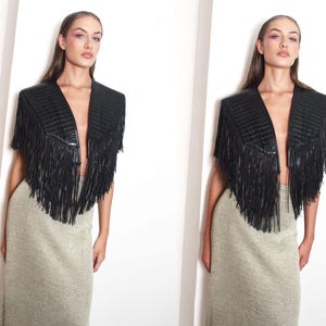May include: A black leather jacket with a fringed design and a V-neck. The jacket features structured shoulders and long fringe detailing. The model is wearing a long, gold-toned skirt.