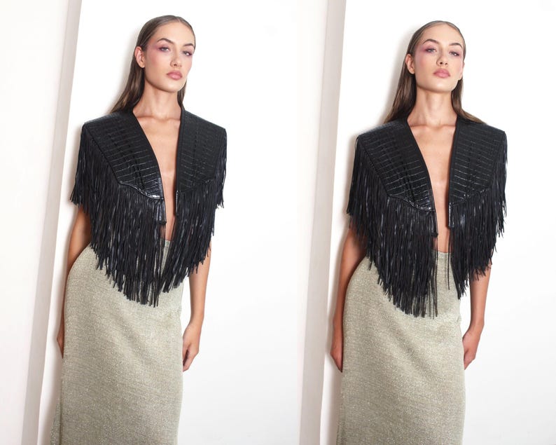 May include: A black leather jacket with a fringed design and a V-neck. The jacket features structured shoulders and long fringe detailing. The model is wearing a long, gold-toned skirt.