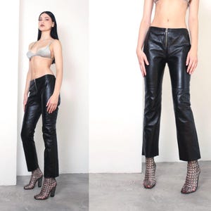 Op de afbeelding: Zwarte leren broek met ritssluiting. De broek heeft een rechte pijp. Het model draagt de broek met een zilveren bh en pumps met patroon. De broek is gemaakt van glanzend leer.