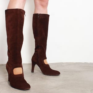 以下が含まれることがあります： サイドにカットアウトのディテールとブロックヒールが付いた、ブラウンのスエードのニーハイブーツのペアです。