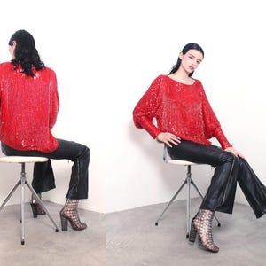 Puede incluir: Un top rojo con lentejuelas, mangas largas y cuello redondo. El top se combina con pantalones de cuero negro y tacones estampados. La modelo está sentada en un taburete blanco sobre un fondo blanco. Un conjunto de moda audaz.