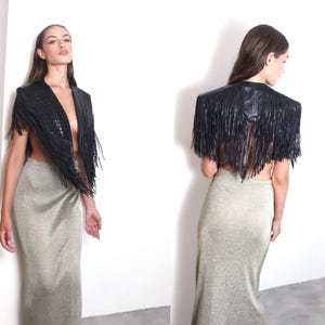May include: A black leather fringed shawl with a textured design. The shawl is worn over a gold-coloured, floor-length skirt. The model is standing against a white wall.