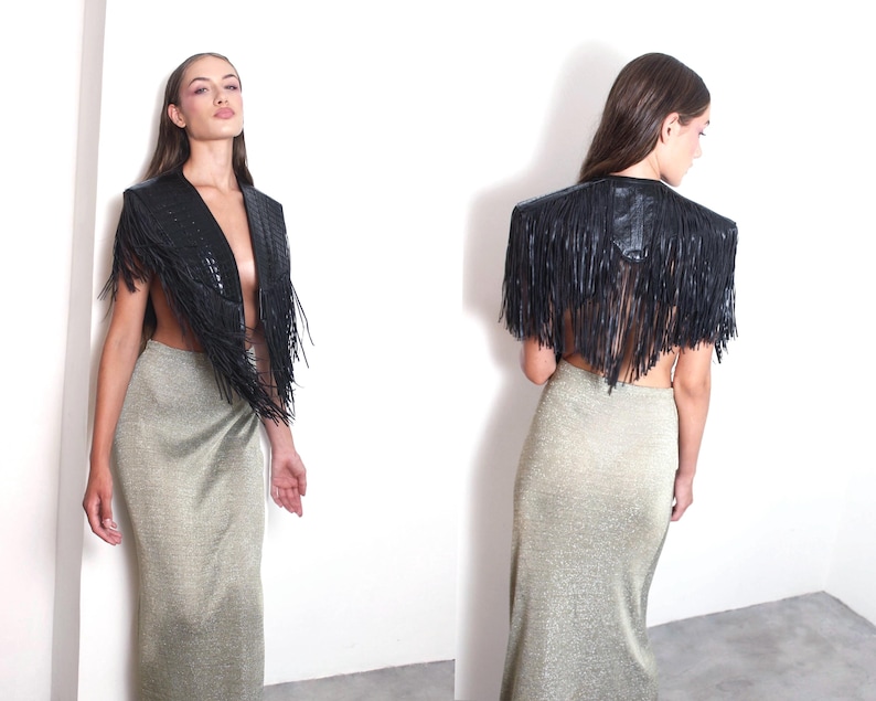 May include: A black leather fringed shawl with a textured design. The shawl is worn over a gold-coloured, floor-length skirt. The model is standing against a white wall.
