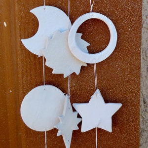 May include: White ceramic moon and star mobile hanging from a brown wall. The mobile features a large crescent moon with a smaller star hanging from it. Other smaller stars, moons, and circles hang from the moon.