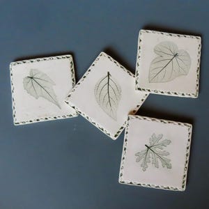 May include: A set of four square white ceramic coasters, each featuring a distinct dark green leaf skeleton print. The edges of the coasters have a textured, dark green speckled border, providing a contrast to the smooth white surface.