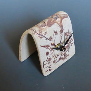 May include: A cream-colored ceramic clock with a curved, open design. The clock face features black hands and a gold-colored center. The surface is decorated with brown botanical illustrations, creating a unique decorative piece.