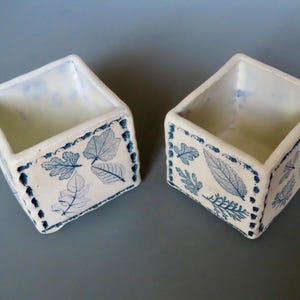 May include: Two square, white ceramic containers with a blue leaf pattern. The containers have a rectangular shape with a slightly flared top. The leaf design is repeated around the sides of the containers.