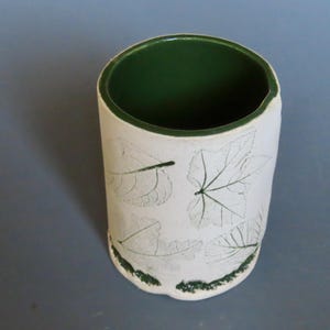 May include: A cylindrical ceramic cup with a white exterior and a green interior. The white surface is decorated with green leaf imprints. The rim and base of the cup are also trimmed with green. The cup is approximately 4 inches tall.