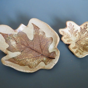 May include: Two ceramic leaf-shaped dishes with brown leaf imprints. The larger dish is a light beige color and the smaller dish is a slightly darker beige.
