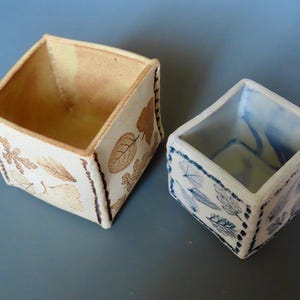 May include: Two square ceramic containers. One is beige with brown leaf designs, the other is white with blue leaf designs. Both have a decorative stitched border. The containers are empty and sit on a gray surface.
