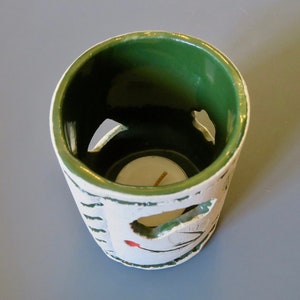 May include: A white ceramic candle holder with a green rim and a cutout design. The holder has a small, round opening in the center for a tea light candle.