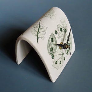 May include: A white ceramic clock with a curved, arch-like design. The clock face features green leaf patterns and black clock hands. The clock measures approximately 6 inches in height.