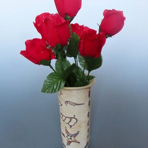 May include: A bouquet of vibrant red roses in a beige ceramic vase. The vase is cylindrical and decorated with brown sea-themed illustrations, including starfish and seahorses. The roses have green leaves and are arranged to create a full, eye-catching display.