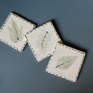 May include: Three square white coasters with a decorative border. Each coaster features a different pressed leaf design in shades of green.