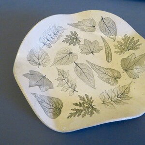 May include: A white ceramic dish with a wavy edge, decorated with a pattern of pressed leaves in shades of green.