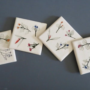 May include: Four square white ceramic coasters with botanical illustrations. Each coaster features delicate drawings of flowers and leaves in shades of red, orange, blue, and pink. The coasters are arranged on a gray surface.