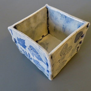 May include: A white ceramic square planter with a blue and white leaf design. The planter has a rough, textured surface.