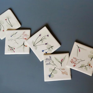 May include: Five white ceramic coasters with pressed flower designs. Each coaster features a different arrangement of dried flowers in shades of red, blue, and white.