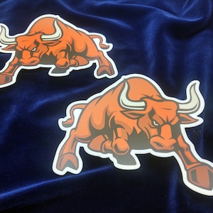 May include: Two orange bull stickers with white horns and a white outline. The bulls are facing forward with their mouths open and their tongues sticking out.