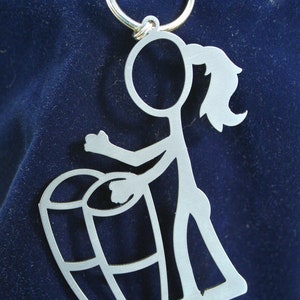 May include: A silver keychain featuring a stick figure playing a drum. The figure is holding a drum with a simple design and has a ponytail.