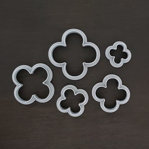 May include: A collection of six gray, clover-shaped cookie cutters of varying sizes. The cutters are made of a light-colored metal and are arranged on a dark brown surface. The clover shapes have four rounded lobes.