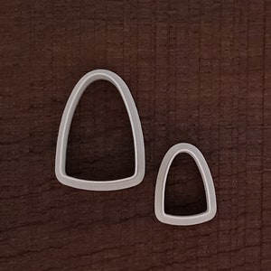 May include: Two white plastic cookie cutters shaped like candy corn. The larger cutter is on the left and the smaller cutter is on the right.