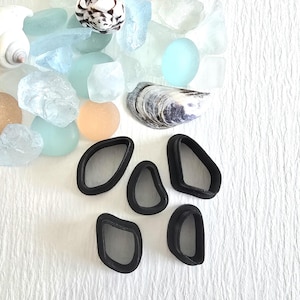 May include: An assortment of sea glass, seashells, and black, geometric-shaped objects. The sea glass is in shades of blue, green, and clear. The seashells are white and tan. The black objects have rounded edges and various shapes.
