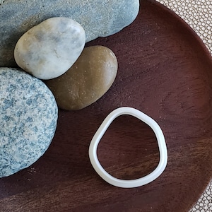 May include: A white, smooth, rounded, ring-shaped cookie cutter on a dark brown wooden surface with four gray and brown stones.