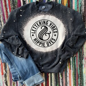 May include: A dark gray sweatshirt with a bleached white circle design. The circle features the text "LETTERING VIBES" and "HIPPIE DYES" in a circular pattern, with a stylized ampersand in the center. Blue jeans are folded next to the sweatshirt.