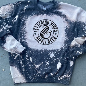 May include: A dark blue sweatshirt with a white bleach splatter design. The sweatshirt has a black circle logo with the text "Lettering Vibes & Hippie Dyes" in white.