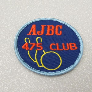 Puede incluir: Un parche bordado azul y blanco con el texto "AJBC 475 CLUB" en rojo y amarillo. El parche presenta un gráfico de bolos y bola.